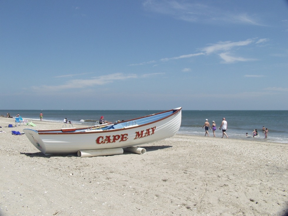 Image of white boat on the beach with the words cape may written on it. There are people on the beach behind the boat.