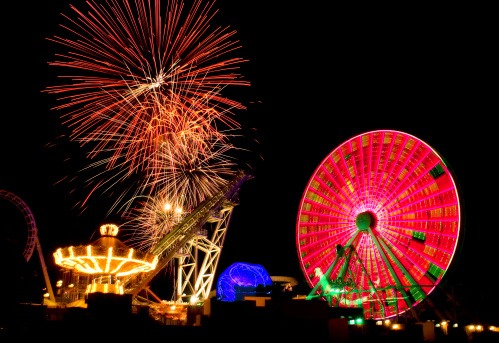 Image of a carousel and other amusement rides with fireworks going off in the night sky in the background