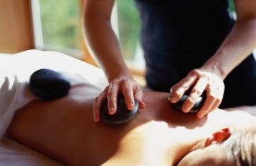 Image of a woman on her stomach with a masseuse placing black hot stones on her back