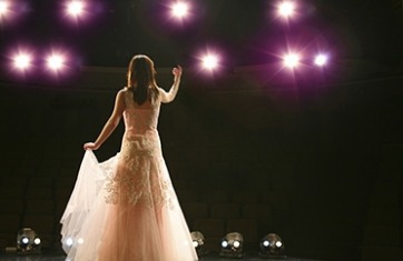 image from behind a woman in a white dress on stage with long brown hair singing to the audience